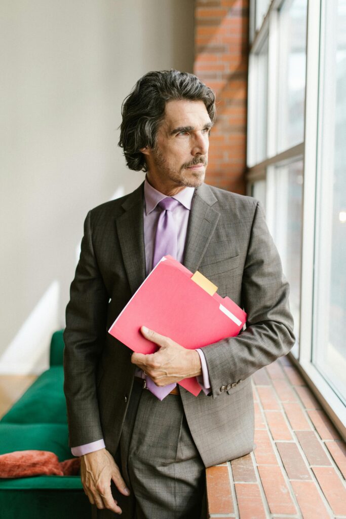 Businessman in suit holding documents, standing by office window. Ideal for corporate themes.